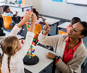 A child with pigtails and wearing a white sweater, builds a tower of blocks with the help of a teacher who has short brown hair, an wears a red blouse and light brown cardigan. Other children are seated at desks in the background.