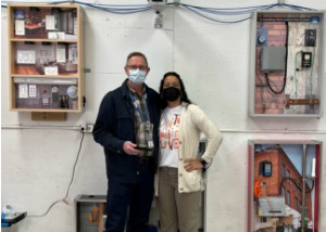 Network Cabling Technician/Smart Home Technology teacher Ed Henson, who has short brown hair and is wearing a protective face mask and eyeglasses, a plaid collared shirt and a dark blue jacket, accepts commitment award from Career & Technical School Principal Shelette Pleat who also wears a protective face mask, and a yellow cardigan over a t-shirt with a graphical design. They are standing together in the Network Cabling classroom and smile for the camera.