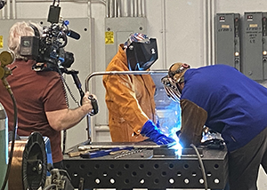 Three people stand around a low metal welding table: a camera person, who is filming the action with a portable video camera, Welding and Metal Fabrication student Francesco DelGallo, who wears a protective welder’s helmet, brown jacket and gloves, and This Old House host Kevin O'Connor, also wearing protective helmet and bright blue jacket. DelGallo and O'Connor together weld metal as blue sparks fly.