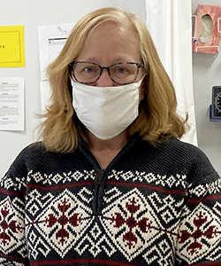 Integrated Science Teacher Joanne Clegg, who has shoulder length reddish-blond hair, and is wearing eyeglasses, a white protective face mask and a red, white and blue nordic-print sweater, looks at and smiles for the camera.