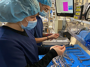 Two Sterile Processing program students, both wearing light blue protective hair coverings and face masks and dark blue scrub shirts, stand side by side and inspect medical tools in a workroom at Albany Medical Center.