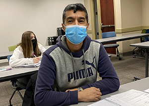Adult learner Zekriya Baz Mohammad, who has short dark hair and wears a bright blue face mask and grey and blue hoodie with the word "Puma" on its front, sits with arms folded on a desk in Capital Region BOCES High School Equivalency classroom and smiles for the camera.