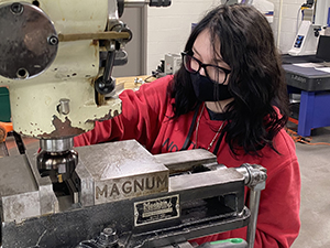 Allison Umstadter, who has long black hair, wears dark rimmed eyeglasses, a black protective face mask and red hoodie, works with a tool in a Career & Technical School manufacturing and machining classroom.
