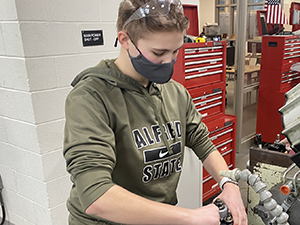 Caitlin Atkins, who has clear protective googles pushed to the top of her head, and is wearing a black protective face mask and an olive green hoodie with the words "Alfred State" printed on it, works at a machine during an on-site experience with a Capital Region business partner.