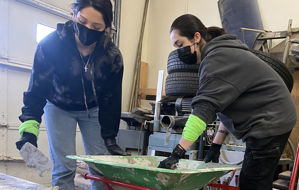 Two young women, both with long black hair pulled back in ponytails, and wearing black, protective facemasks and dark colored hoodies, use tools to scoop mortar from a wheelbarrow and build a brick wall in a Career & Technical School Construction Trades classroom.