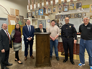 Student Daniel Baughn, who has short brown hair, eyeglasses and is wearing a tan long-sleeved sweater and jean, stand beside a podium constructed for use by City of Watervliet representative. Baughn is standing with Watervliet City, school and BOCES officials and teachers in a Career and Technical School Building Trades classroom.  