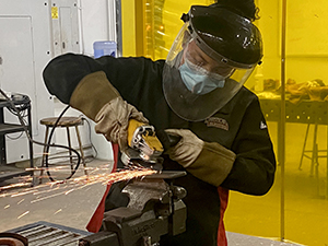 Darielys Maya, who has dark hair pulled back in a curly ponytail and is wearing a welder's hood and clear face shield, gloves and jacket, used a tool to grind metal held in a vise in a Career and Technical School classroom. 