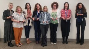 Eight members of the Capital Region BOCES Instructional Coaching team stand in a row, holding individual commitment awards for excellence and smiling for the camera.