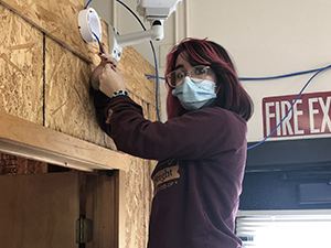 Kira St. Andrews, who has long black hair streaked with pink, and is wearing round eyeglasses, a light blue face mask and purple hoodie, works on wiring attached to plywood wall in a Career & Technical School Network Cabling classroom.