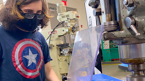 Aidan Matile, who has shoulder length brown hair, and wears black eyeglasses, a black face mask and blue and red t-shirt with a Captain America shield on its front, works with equipment in a Career & Technical School Manufacturing and Machining classroom.
