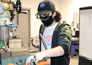  Aidan Matile, who has shoulder length brown hair, and wears a black ball cap with a NASA patch on its front,  black eyeglasses, a black face mask, a grey NASA t-shirt and camo print hoodie, works with equipment in a Career & Technical School Manufacturing and Machining classroom.