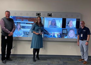 The Northeastern Regional Information Center’s Supervisor of Instructional Resources Mike Sylofski, Instructional Technology Service Manager Lora Parks and Assistant Superintendent Michael Doughty stand in a row in front of wall mounted display screen featuring five members of the Cooperative Virtual Learning Academy. All are smiling for the camera. 