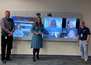 The Northeastern Regional Information Center’s Supervisor of Instructional Resources Mike Sylofski, Instructional Technology Service Manager Lora Parks and Assistant Superintendent Michael Doughty stand in a row in front of wall mounted display screen featuring five members of the Cooperative Virtual Learning Academy. All are smiling for the camera.