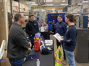 Two adult men representing area businesses and two Heating, Ventilation, Air Conditioning and Refrigeration (HVAC/R)students talk while standing around a long table in a career and technical school garage.