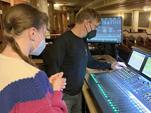 Entertainment Technology student Chloe Hollner-Turner, who has light brown hair pulled into a ponytail and wears a blue face mask and white, blue and pink diagonally striped sweater, stands alongside a technician behind a soundboard at Proctors Theatre in Schenectady, N.Y.