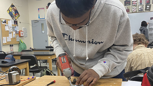 Student Dharma Parmeshwari, who has short dark hair and is wearing black eyeglasses and a light grey hoodie with the word Champion on its front, looks down while using a hand drill at a workbench in a Heating, Ventilation, Air Conditioning and Refrigeration classroom