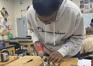 Student Dharma Parmeshwari, who has short dark hair and is wearing black eyeglasses and a light grey hoodie with the word Champion on its front, looks down while using a hand drill at a workbench in a Heating, Ventilation, Air Conditioning and Refrigeration classroom