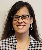 Assistant Principal Kristen Roys, who has long black hair and is wearing dark rimmed eyeglasses, dangly silver earrings and a black and white checked collared shirt, looks at and smiles for the camera.