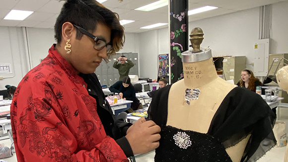 Global Fashion student Robert Vincent, who has short dark hair with a longer bang, and wears black eyeglasses, a long gold earring and printed red shirt, sews the sleeve of a black formal dress that is displayed on a mannequin.