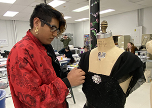 Global Fashion student Robert Vincent, who has short dark hair with a longer bang, and wears black eyeglasses, a long gold earring and printed red shirt, sews the sleeve of a black formal dress that is displayed on a mannequin. 