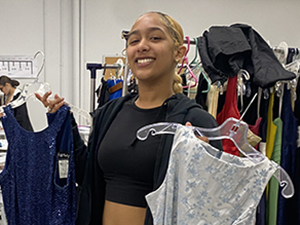 A Global Fashion student, who has blond hair pulled back in a low ponytail, and is wearing a black hoodie and cropped top, stands in front of a rack of colorful dresses holding a blue and a white prom dress in each hand and smiling for the camera.