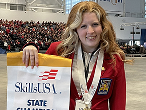 Global Fashion student Jocelyn Howe, who has long wavy blond hair and is wearing a red blazer and white collared shirt, stands in an auditorium with a crowd seated in the bleacher at the rear. Jocelyn is holding a banner that reads "SkillsUSA State Championship Job Interview" and is wearing a first place medal.