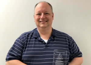 Health Safety Risk Program Coordinator Michael Walker, who is wearing a dark blue and white striped polo and holding a translucent commitment award, looks at and smiles for the camera. 