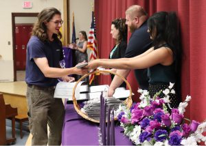 A smiling electrical trades student, who has shoulder length brown hair and mustache, and is wearing eyeglasses, a dark blue t-shirt and khaki pants, shakes hands with a teacher across a table draped in purple. There is a basket of National Technical Honor Society drapes, pins, certificates, a flower arrangement in purple and white and a set of tall purple candles on the table, which is set on a stage.
