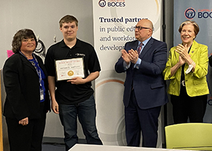 Construction/Heavy Equipment senior Nate Bartlett, who has short brown hair and is wearing a dark blue polo shirt and blue pants, holds a scholarship certificate and smiles for the camera. From left to right, Capital Region BOCES' Nancy Liddle and Senior Executive Officer Joseph Dragone, and New York State Department of Labor Commissioner Roberta Reardon stand with Nate and cheer this accomplishment. 