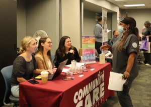  An adult nursing student, who has long brown braids and is wearing a black head band, a light blue protective face mask and dark grey nursing scrubs, stands speaking with three people who are seated at a table draped in a red cloth that displays the words "Community Care."
