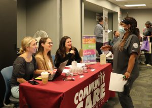An adult nursing student, who has long brown braids and is wearing a black head band, a light blue protective face mask and dark grey nursing scrubs, stands speaking with three people who are seated at a table draped in a red cloth that displays the words "Community Care."