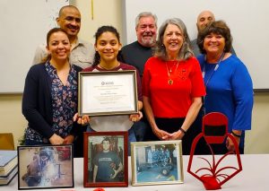 Welding student Darielys Maya, who is holding a framed scholarship certificate, stands surrounded by family, Evan Schwartz' parents and Career and Technical School teachers. Maya is the recipient of the Evan M. Schwarz Helping Hand Memorial Scholarship. All the people pictured are standing and smiling for the camera. A collection of photos of Evan Schwartz are displayed on a table in front of the group. 