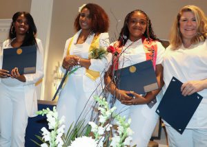 A group of four graduates of the Capital Region BOCES Adult Practical Nursing program stand side by side. All are wearing white clothing and holding blue graduation certificates. They are all looking at and smiling for the camera. 