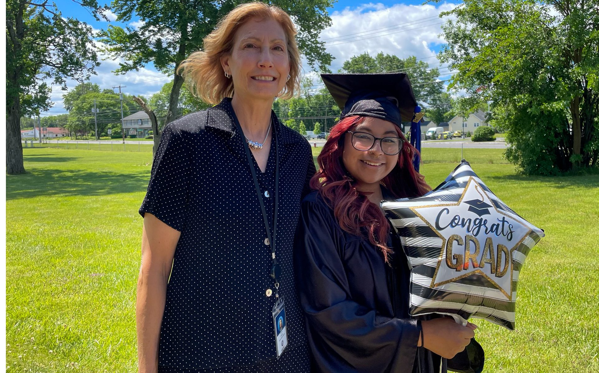 A Capital Region BOCES teacher, who has blond hair and is wearing a black dress with small white polka dots, stands with an arm around a graduate of our Maywood school program. The student is wearing a black graduate's cap and gown, has long, wavy bright red hair, wears black eyeglasses and is holding a star-shaped white and black striped helium ballon with the words "Congrats Grad." They are outside on a bright, sunny day and are looking at and smiling for the camera.