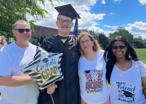A graduating student, dressed in a black cap and gown and holding a black and white star-shaped helium balloon with the words "Congrats Grad" stands outside on a sunny day with family members on the left and right. All are looking at and smiling for the camera.