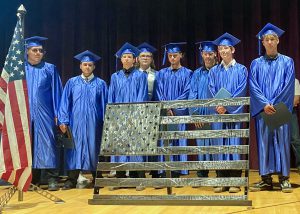 A row of welding students, wearing bright blue graduate caps and gowns, stand in a row behind a grey steel sculpture of the American flag, which they made as a gift to Capital Region BOCES. They are all looking forward and smiling for the camera. A red, white and blue American flag in a stand shows at the left of the photo.
