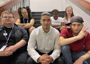 Six students sit in two rows on a staircase. They are all dressed for their program graduation ceremony. All look at and smile for the camera. One student, at the right, holds their hand in a peace sign. 