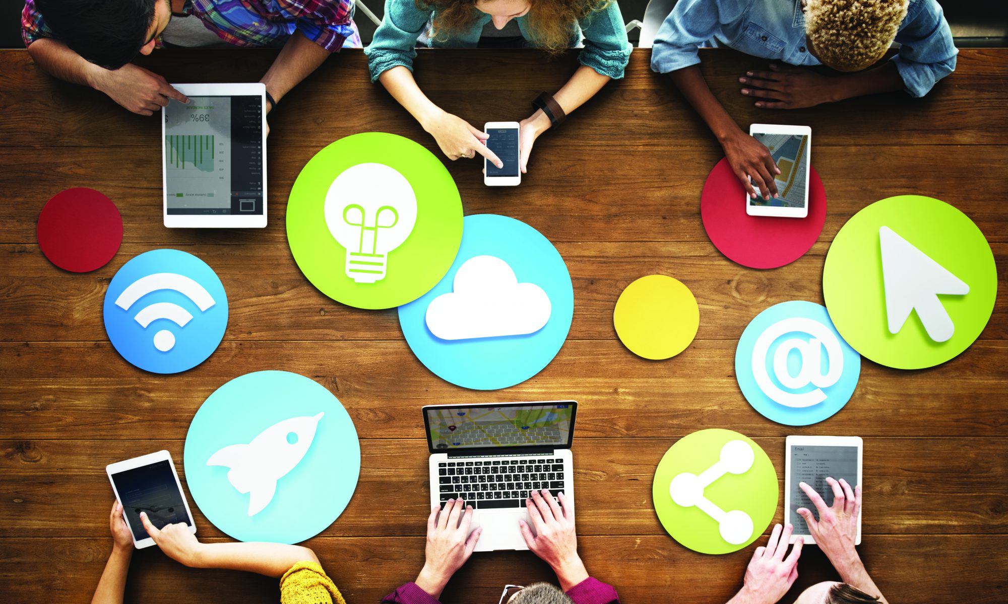 A diverse group of young adults, each interacting with different electronic devices, sit on either side of a wood-topped table. Bright red, blue and green circles with various white icons that represent different forms of communication are featured in the center of the table.