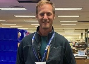 May 2022 commitment award recipient John Florussen, who has short, reddish blonde hair, and is wearing a grey fleece jacket, stands in front of Florussen is looking at and smiling for the camera.