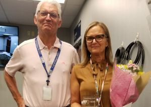 June 2022 commitment award recipient Sue Frank, who has long, light brown hair, and is wearing cats-eye shaped glasses and a gold colored blouse, stands beside Shared Food Services Director Jeff Bradt, who has short white hair, wears eyeglasses and a white polo shirt. Both are looking at and smiling for the camera.