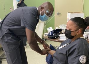 Adult Practical NUrsing graduate Tenniel Thompson, who has a shaved head and is wearing a clear, protective face shield and light blue face mask, leans in to take the pulse of seated classmate. They are together in an Adult Practical Nursing classroom. Both are wearing dark blue nursing scrubs.