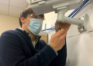 Network Cabling Technician/Smart Home Technology program graduate, Thomas Rodick, who has short light brown hair and is wearing clear protective eyewear, a light blue face mask and a collared dark blue sweater, looks at and uses a tool to work on equiptment in a Career & Technical School classroom.