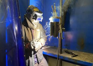 An adult learners, wearing a protective welders' helmet and clothing, uses a tool to weld metal. The student is standing in an enclosed space with a partially drawn clear curtain and a bright blue background.