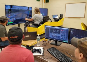 Heavy Equipment Teacher Andrea Rea sits facing two large screens that show a simulation of the action of a piece of construction equipment. Three others are seated in the foreground at a table, where a computer keyboard and screen are placed.