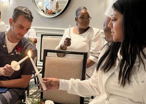 Adult Practical Nursing graduate Chris Davis, who has short brown hair and is wearing dark blue nursing scrubs with a red rose boutonniere, holds a candle toward another graduating student's lit candle as they take part in a graduation ceremony. Both graduates are seated and another graduate, also holding a candle, is seated behind them.