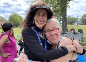 Training Specialist Donna Lamkin, who has long wavy dark brown hair and is wearing a black sun visor and long sleeved black top, hugs Michelle Rosenberger, who has greying blond hair and wears red eyeglasses. They are outdoors on the front lawn of the Maywood School. They are standing in front of rows of folding chairs and are facing and smiling for the camera.