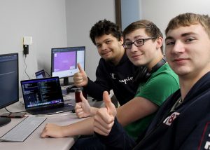 A group of three students sit in a row, facing toward a table at left on which numerous computer keyboards and monitors are placed. They are looking at and smiling for the camera and giving a thumbs up. 