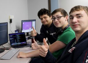 A group of three students sit in a row, facing toward a table at left on which numerous computer keyboards and monitors are placed. They are looking at and smiling for the camera and giving a thumbs up.
