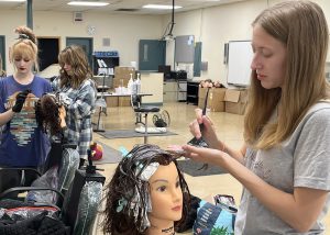 Madison Scheur, who is featured standing at the right side of the image taken in a cosmetology classroom, uses a color brush to apply hair color to a mannequin head. Madison has long light brown hair and is wearing a light grey t-shirt.