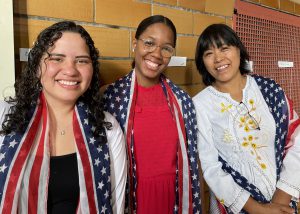 Three adult students who are women stand in a row, in front of a  brown tiled wall. All are wearing American flag-printed scarves over their shoulders, and are looking at and smiling for the camera.